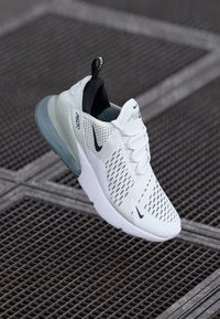 Bijela i siva Nike Air Max 270 tenisica s crnim swoosh logotipima, mrežastim gornjim dijelom i vidljivim zračnim jastučićem na pozadini od metalne rešetke.