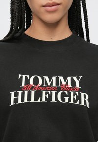 Μαύρο φούτερ με στρογγυλή λαιμόκοψη, με μεγάλο λευκό κείμενο "TOMMY HILFIGER" και κόκκινη καλλιγραφική επιγραφή "All American Classics" στο μπροστινό μέρος.