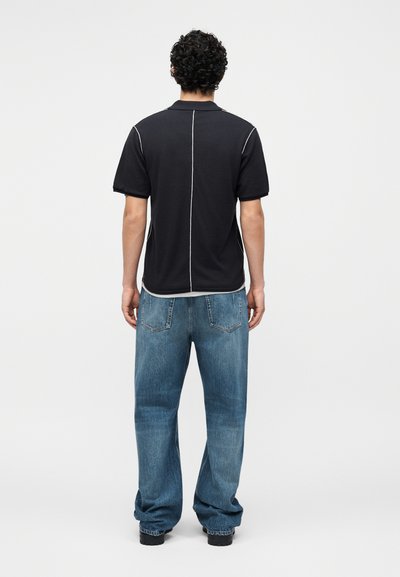 Άνδρας με κατσαρά μαλλιά που φοράει μαύρο πόλο με κοντά μανίκια και λευκές ραφές καθώς και φαρδιά μπλε τζιν, στέκεται γυρισμένος με την πλάτη σε μονόχρωμο φόντο.