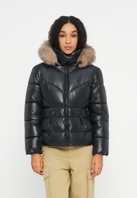 Frau in einer schwarzen, gesteppte Daunenjacke mit einem Kapuzenrand aus Pelz und beigen Cargo-Hosen, die vor einem schlichten weißen Hintergrund steht.
