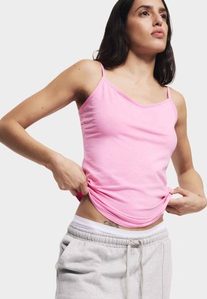 Camisola di cotone rosa con spalline sottili, design a V, e texture liscia. Indossata con pantaloni della tuta grigi, che mostrano un look casual.