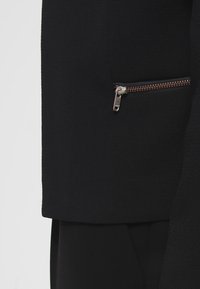 Černá bunda se zipovou kapsou a kovovými detaily, hladká textura a strukturovaný design. Čisté linie bez viditelných vzorů.