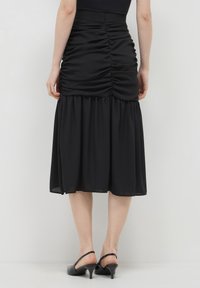 Eine Person trägt einen schwarzen, gerafften Midirock und schwarze Slingback-Absätze, von hinten vor einem schlichten weißen Hintergrund gezeigt.