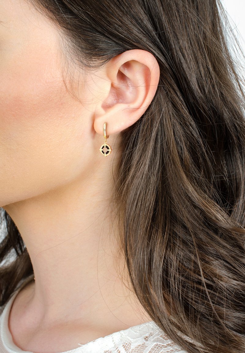 Orecchini a cerchio d'oro con un design geometrico e un particolare centrale nero. Il modello ha lunghi capelli ondulati e una pelle liscia.
