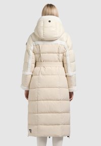 Lange crèmekleurige donzen jas met een tailleceintuur, een zachte voering en bontaccenten aan de capuchon en manchetten. Voorzien van een logo linksonder.