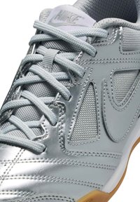 Sapatilha desportiva prateada com um upper lustroso e elegante, com detalhes em malha, atacadores finos e uma sola de borracha de gingim. Logótipo da Nike visível na língua.