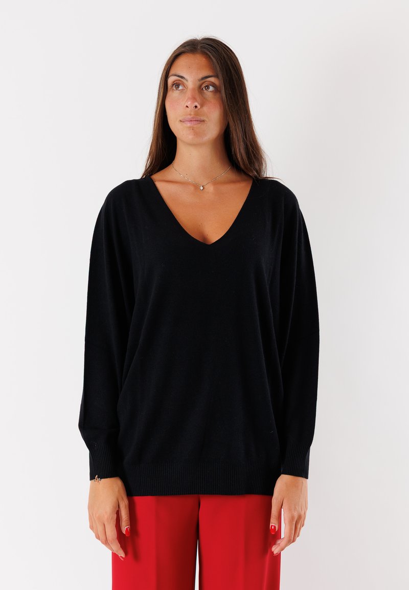 Donna con lunghi capelli castani, che indossa un maglione nero a V e pantaloni rossi, in piedi contro uno sfondo bianco, guardando leggermente a destra.