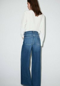 Jeans anchos de mezclilla azul con cintura alta, que cuentan con dos bolsillos traseros y un dobladillo limpio. Combinados con una blusa blanca de manga larga.