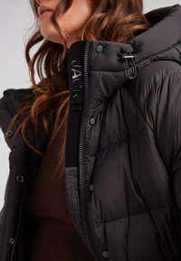 Kobieta w czarnej kurtce puchowej z kapturem i widocznymi detalami zamka, częściowo zapięta na brązowej bluzie, z falującymi włosami.
