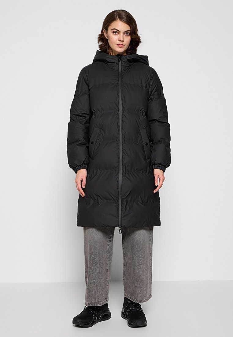 Manteau rembourré noir avec capuche, fermeture éclair et poches latérales. Présente une texture matelassée et une longueur jusqu'aux genoux. Porté par-dessus un pantalon gris.