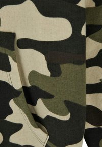 Tissu à motif camouflage dans des teintes de vert et de beige, présentant une texture douce et une poche visible. Le design comprend des formes irrégulières et fluides.