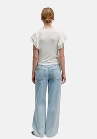 Weißes Kurzarmoberteil mit gerafften Schultern, kombiniert mit hellblauen weit geschnittenen Jeans. Der Stoff ist glatt, und die Jeans verfügen über zwei Gesäßtaschen.