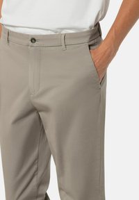 Mann mit beigen Hosen und weißem Hemd, die rechte Hand in der Tasche, zeigt einen Knopf und die Gürtelschlaufen der Hose.