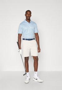 Ljusblå pikétröja med mörka prickar, korta beige shorts, marinblå bälte, vita sneakers och golfklubba, presenterad mot en enfärgad bakgrund.