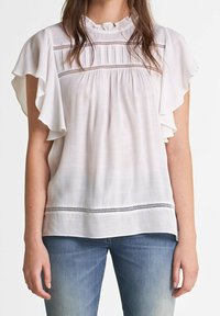 Blouse blanche à manches volantées, avec un col plissé et un détail en dentelle sur la poitrine. Portée avec un jean en denim bleu.