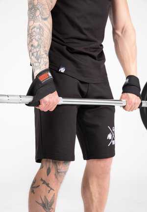 Getatoeëerde persoon met zwarte sporthandschoenen en kleding grijpt een halterstang vast en bereidt zich voor om gewichten te tillen.