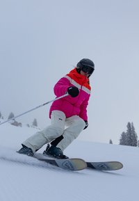 Attrezzatura da sci con giacca rosa brillante e bianca, pantaloni bianchi, guanti neri e casco da sci. Gli sci sono neri con un design distintivo. Sfondo innevato.