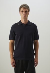 Polo shirt marine avec une finition texturée, manches courtes et un col or à liseré contrastant ; comporte une patte de boutonnage à trois boutons et une étiquette sur le côté.