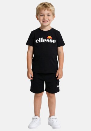 Jeune garçon souriant portant un t-shirt et un short noirs ellesse, debout avec des baskets blanches sur un fond clair uni.