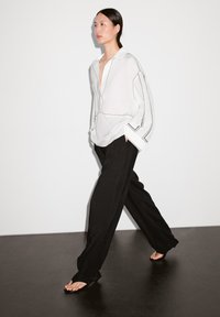 Chemise blanche décontractée avec des surpiqûres noires contrastées, associée à un pantalon large noir et des sandales plates. Tissu lisse, coupe décontractée.