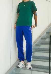 Vihreä puuvillapaita, jossa on logo, siniset polyesteriset urheiluhousut valkoisilla raidoilla ja moniväriset urheilukengät yksityiskohdilla.