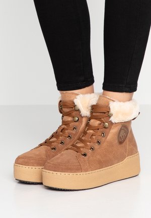 Brązowe botki na platformie z zamszu z wiązaniem z przodu i białą futrzaną podszewką, noszone z czarnymi obcisłymi spodniami.