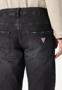 Temno sivi jeans s srednjim pasom, ki imajo dva zadnja žepa in trikotno rdečo oznako na desnem žepu.