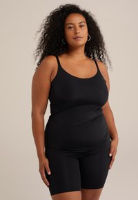 Ženska s kodrastimi lasmi, oblečena v črno brezrokavno majico in usklajene črne kratke hlače, stoji pred bež ozadjem.