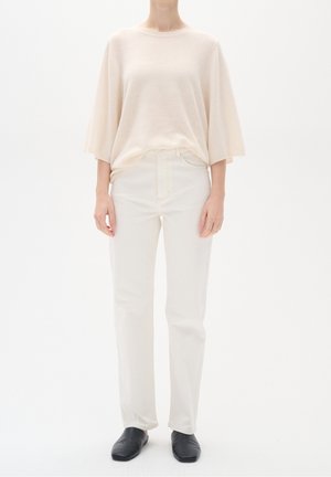Lichtbeige, oversized gebreide trui met korte mouwen, gecombineerd met witte rechte jeans en zwarte instappers. Eenvoudig, minimalistisch ontwerp.