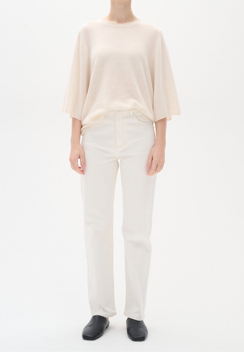 Suéter de punto de color beige claro y de gran tamaño, con mangas cortas, combinado con jeans blancos de pierna recta y zapatos negros sin cordones. Diseño simple y minimalista.