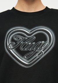 Μαύρο φούτερ με σχέδιο καρδιάς σε λευκή διαβάθμιση και τη λέξη "Frugo" σε καλλιγραφική γραφή στο κέντρο στο στήθος.