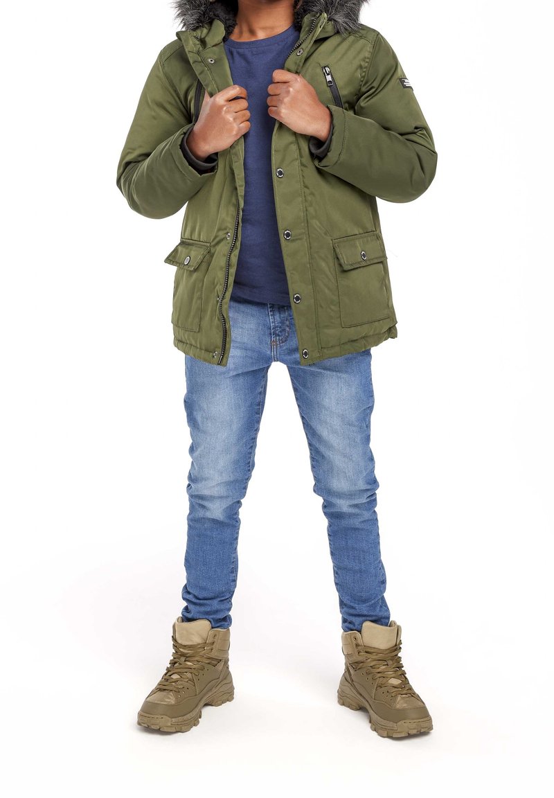 Olivgrüne Jacke mit einer mit Fell besetzten Kapuze, Reißverschluss und Knopfverschluss, zwei Fronttaschen. Getragen über einem marineblauen Hemd, kombiniert mit blauen Jeans und beige Stiefeln.