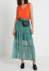 Orange ærmeløs top med en V-hals, kombineret med et grønt blomsterprintet maxiskørt. Outfittet inkluderer en sort bæltepose og spidse sorte flade sko.