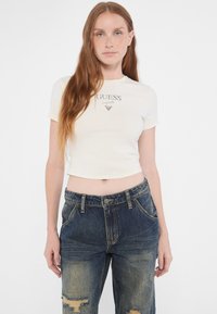 Crop top corto blanco de manga corta con el logotipo "GUESS" en plata, combinado con pantalones vaqueros anchos desgastados de cintura alta en azul oscuro, con costuras visibles.