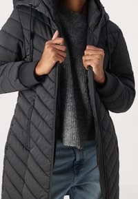 Abrigo acolchado negro con capucha, con cierre frontal y puños elásticos, superpuesto a un suéter de punto gris y unos jeans azules.