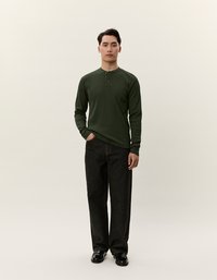 Sötétzöld, hosszú ujjú henley ing gombos gallérral, széles lábú fekete farmernadrággal párosítva. A modell egy semleges háttér előtt áll.