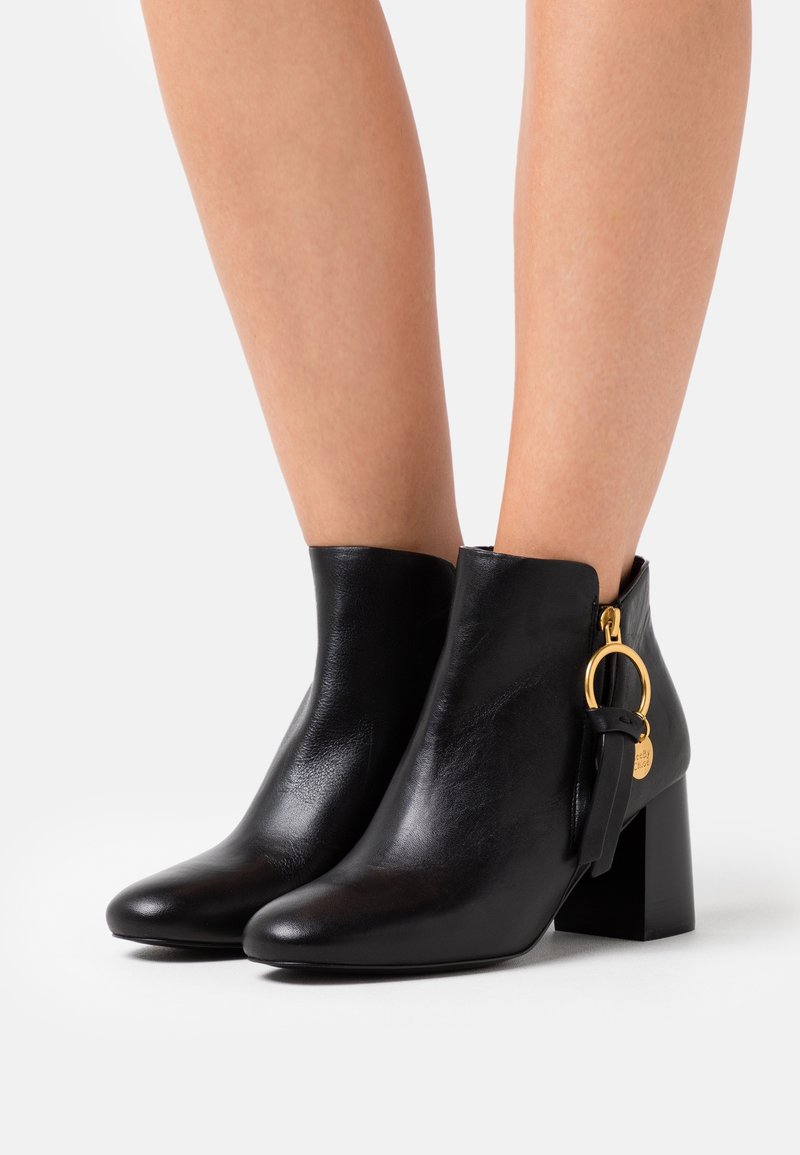 Bottines en cuir noir à talons carrés avec tirettes de fermeture éclair dorées en forme de cercle, portées sur des jambes nues sur un fond blanc.