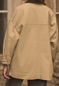 Beige ylättävä takki, joka on valmistettu puuvillasta ja jossa on vakosuora kaulus. Ominaisuuksia leveät hihat ja näkyvät ompeluyksityiskohdat. Yksinkertainen takakuvio.