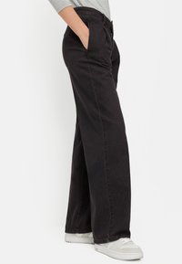 Schwarze Hosen mit weitem Bein, glatter Textur und vorderen Taschen. Verfügen über einen Knopfverschluss und eine lässige Passform. Kombiniert mit weißen Sneakern.