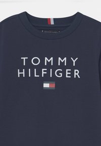 Maglietta di cotone blu navy con la scritta "TOMMY HILFIGER" in bianco, con un piccolo logo rosso, bianco e blu vicino all'orlo.