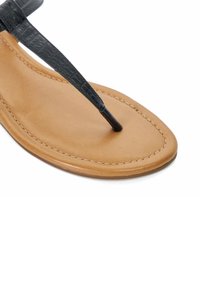 Zwarte faux leren slipper met banden over een lichtbruine platte zool met stiksels, waarmee een gladde textuur en een minimalistisch ontwerp worden getoond.