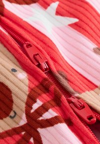 Röd zip-up kläder med ribbad textur, med djur- och stjärnmönster i nyanser av rosa och brunt mot en livfull bakgrund.