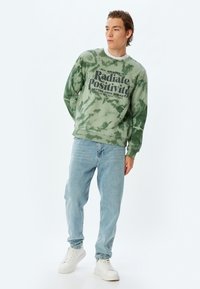 Grön tie-dye sweatshirt med texten "Sprid Positivitet", tillsammans med ljusblå jeans och vita sneakers. Mjuk tygkvalitet och avslappnad passform.
