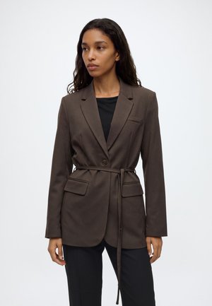 Femme aux longs cheveux bouclés portant un blazer marron ceinturé sur un top noir et un pantalon noir, debout devant un fond blanc uni.