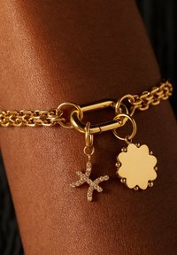 Gouden kettingarmband met een gestructureerd schakelontwerp. Twee bedels: een zeester met fonkelende accenten en een gladde, bloembolvormige tag.