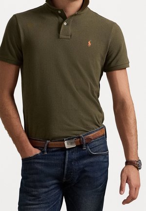 Man die een olijfgroen poloshirt met oranje logo, blauwe jeans, een bruine leren riem en een bruine polshorloge draagt, staand met één hand in de zak.