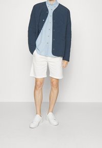 Navy kurtka pikowana, jasnoniebieska koszula na guziki, białe szorty i białe trampki. Gładkie tekstury z widocznymi szwami i detalami guzików.