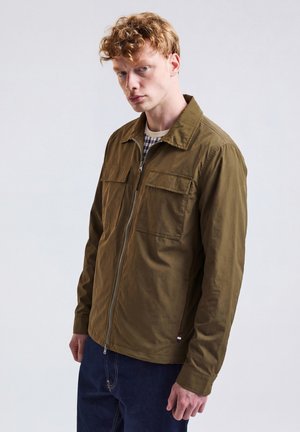 Junger Mann mit lockigem roten Haar, der eine olivgrüne Jacke mit Reißverschluss über einem karierten Hemd trägt, stehend vor einem schlichten hellen Hintergrund.