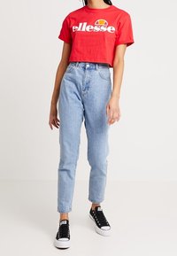 Camiseta corta roja con logo blanco de "ellesse", jeans de tiro alto de color azul claro y zapatillas negras con suelas blancas. Diseño y ajuste simples.