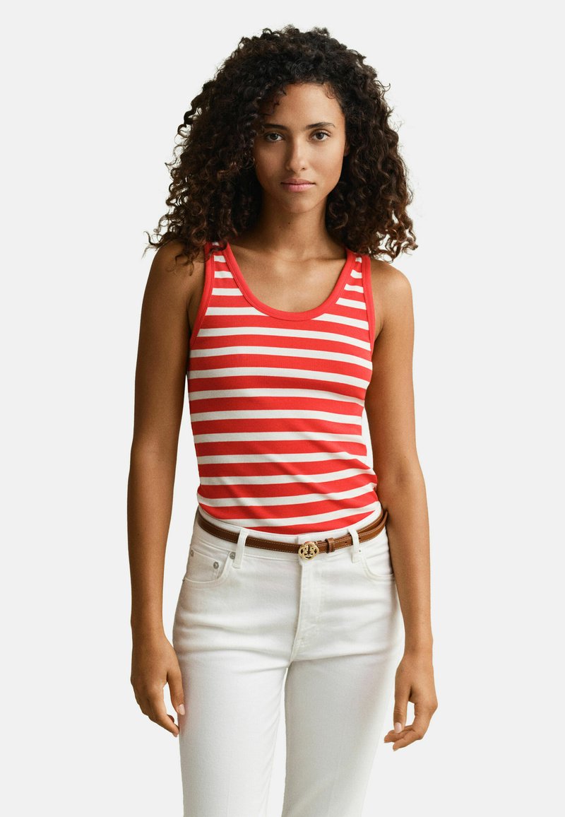 Femme aux cheveux bouclés portant un haut sans manches rouge et blanc à rayures, rentré dans un pantalon blanc avec une ceinture marron, debout devant un fond uni.